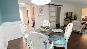 Un coin repas avec une table à plateau en verre, des chaises blanches avec des coussins bleus, un luminaire suspendu et une affiche accrochée au mur.
