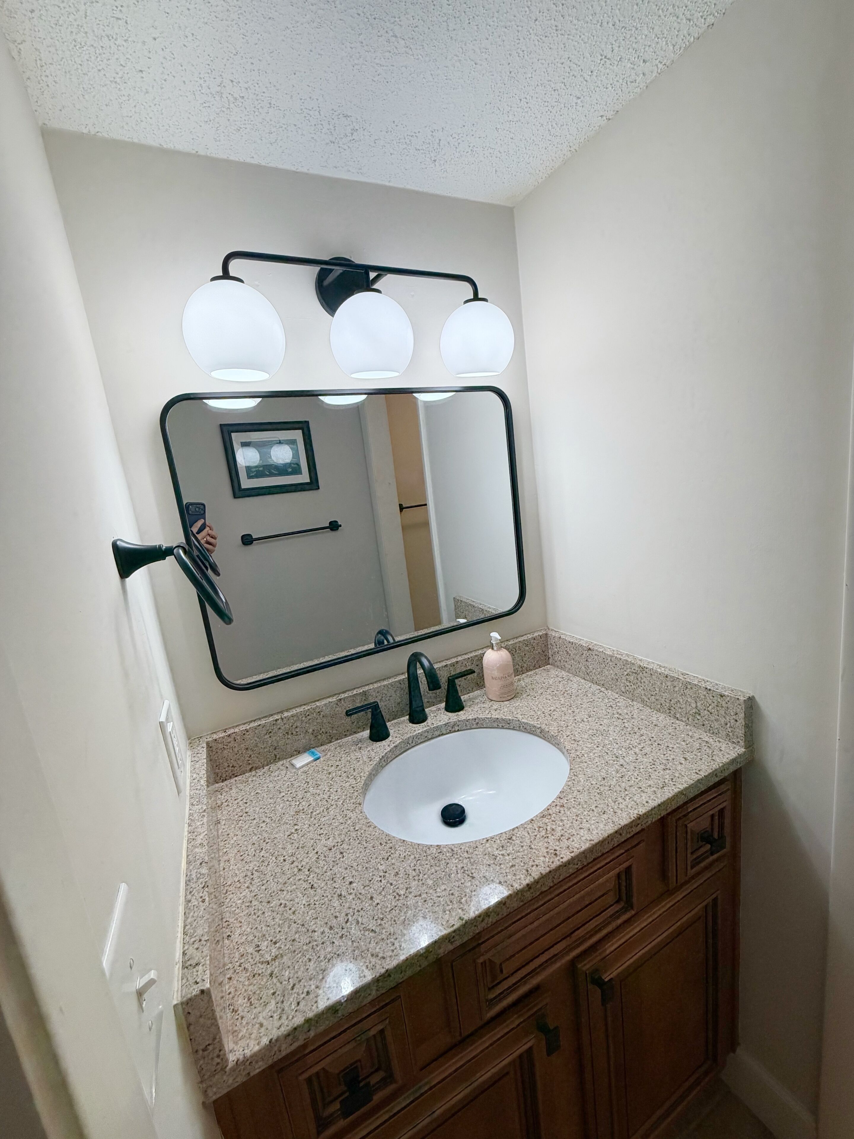 A bathroom with a granite countertop, a white sink, a mirror with lights, and wooden cabinets.