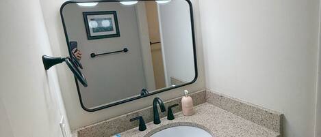 A bathroom with a granite countertop, a white sink, a mirror with lights, and wooden cabinets.