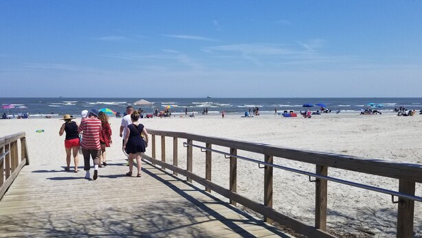 Une promenade en bois mène à une plage de sable où l’on trouve des parasols et des personnes qui se prélassent au soleil.