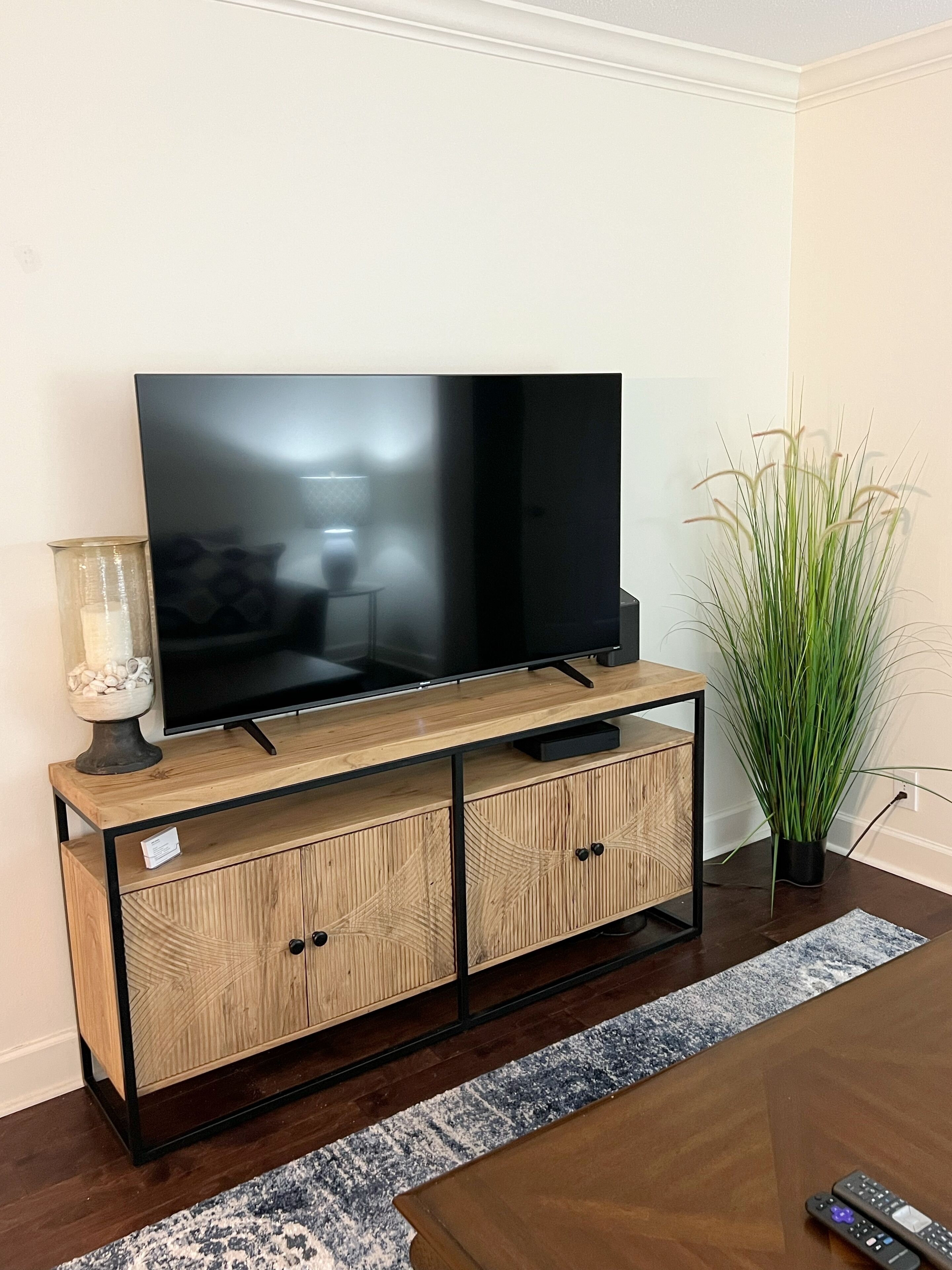 A flat-screen TV on a wooden TV stand with two cabinets, a glass vase with decorative items, and a tall green plant.