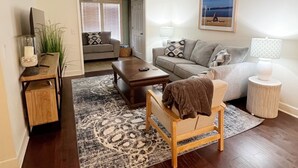Un salon avec un canapé, une table basse et une petite chaise en bois. Il y a un tapis sur le sol et une lampe sur une table d’appoint.