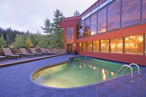 Un edificio moderno con piscina, sillas de descanso y un fondo de bosque.