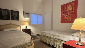 Un dormitorio con dos camas, un ventilador de techo, una ventana con persianas, una mesita de noche roja y una pared con una obra de arte floral.