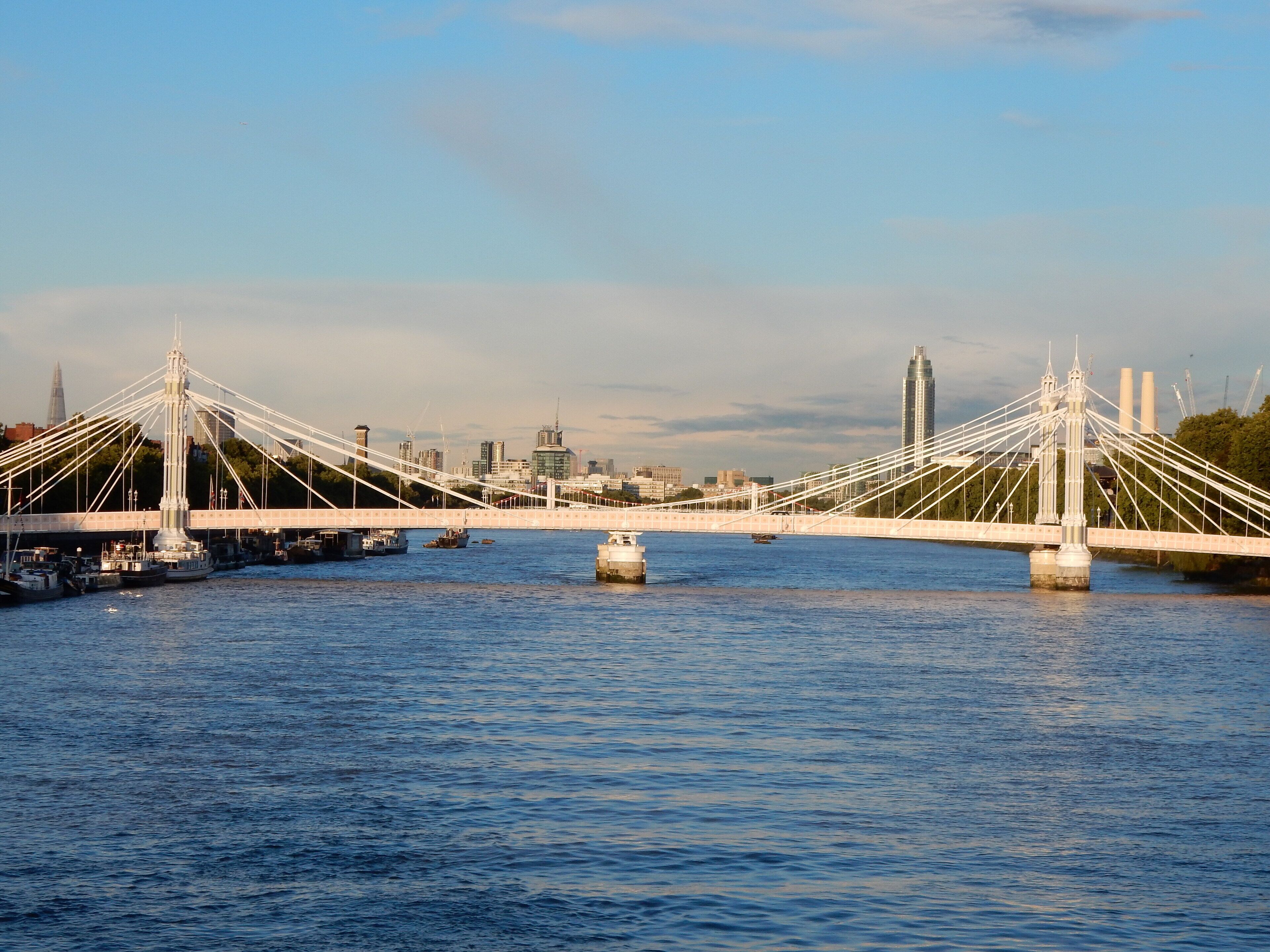 A suspension bridge over a river with boats and a city skyline in the background.