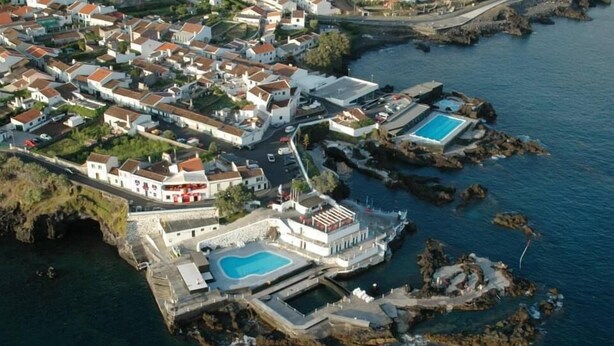 Une vue aérienne d’une ville côtière, où l’on aperçoit un imposant bâtiment blanc doté d’une grande piscine et d’une piscine plus petite, le tout entouré d’un paysage rocheux et bordé par une mer d’un bleu limpide.