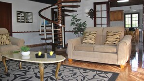 Un salon avec un escalier en colimaçon, un canapé beige agrémenté de coussins à motifs, une table basse ornée d’objets décoratifs et une plante en pot.