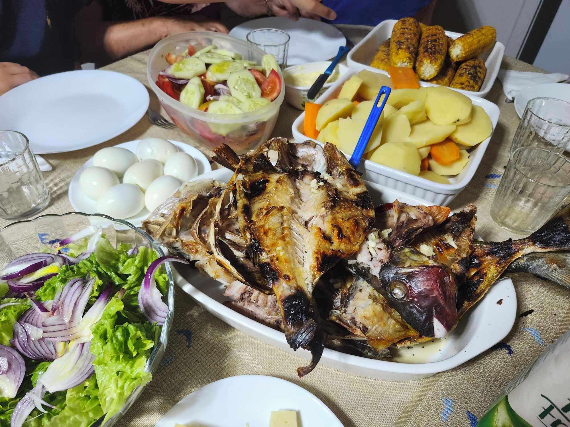 Una mesa con pescado asado, ensalada, huevos cocidos, elote y papas.