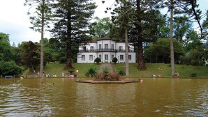 Un grand bâtiment blanc doté d’un balcon, entouré d’arbres et bordé par un plan d’eau.