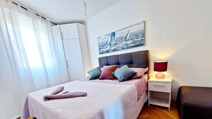 A neatly made bed with a pink bedspread and pillows, a bedside table with a lamp, and a painting of sailboats on the wall. - Aquamarine beach apartment- terrace, seaside (Split)