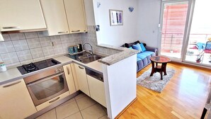 A compact kitchen with a gas stove, oven, and sink, a small dining area with a table and chairs, and a sofa in a living room with a view through a sliding glass door. - Aquamarine beach apartment- terrace, seaside (Split)