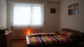 A bedroom with a bed, a radiator, a window with curtains, a desk, and a painting on the wall.