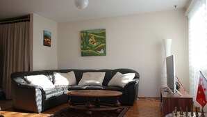 A living room with a black leather sofa, a wooden coffee table, a painting on the wall, a flag, and a television on a wooden stand.