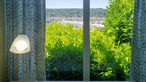 A room with a bed, a lamp, and a window with a view of greenery and a distant landscape. - Poulsbo Marina and Olympic Hideaway View (Poulsbo)