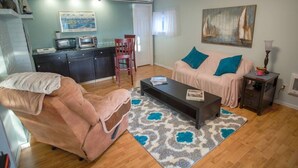 A living room with a sofa, a coffee table, a pink chair, a small kitchen area with a microwave, and a painting on the wall. - Poulsbo Marina and Olympic Hideaway View (Poulsbo)