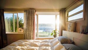 A bedroom with a large bed, a window with curtains, a plant on the windowsill, and a shelf with decorative items. - Poulsbo Marina and Olympic Hideaway View (Poulsbo)