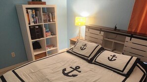 A bedroom with a bed featuring anchor-themed bedding, a bookshelf, a lamp, and a painting on the wall.