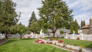 Une pelouse bien entretenue, typique d’une zone résidentielle, avec un jardin central, entourée d’une clôture blanche en bois et des maisons en arrière-plan.