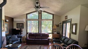 A living room with a large window, a ceiling fan, a fireplace, and a dining area. - A wonderful, secluded 2 story cabin, deck, fire pit, stocked pond for fishing.   (Paradise)
