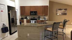 A modern kitchen with granite countertops, wooden cabinets, a stainless steel refrigerator, and a microwave. There are two bar stools with cushioned seats. - Beautiful Home near Walt Disney World   (Davenport)