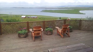 Terraza de madera con dos sillas y macetas, con vista a un cuerpo de agua y campos verdes.