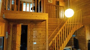 Interior de una cabaña de madera con una escalera central, un balcón y una lámpara colgante.