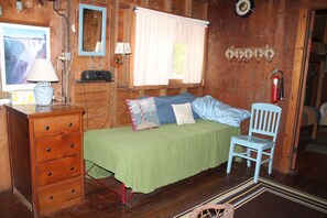 Une chambre dans une cabane en bois, comprenant un lit, une commode, une chaise, une lampe, une fenêtre avec des rideaux et un extincteur.