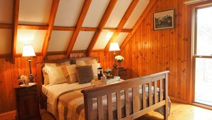 Ein gemütliches Schlafzimmer mit schrägem Dach, Holzwänden, einem Bett mit vielen Kissen, Nachttischlampen und einem Fenster mit Blick.