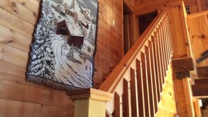 Interior de una cabaña de madera con una escalera y un cuadro de un paisaje nevado.
