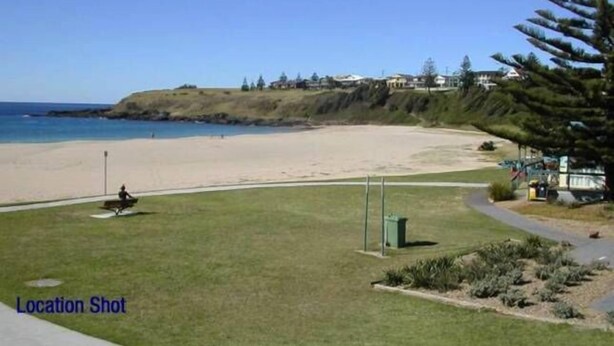 A beach view with a grassy area, a pathway, and a few buildings in the background. - Beach Break - located at Kiama (Kiama)