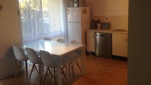 A compact kitchen with a dining table and chairs, a refrigerator, a dishwasher, and a small window with curtains. - Beach Break - located at Kiama (Kiama)