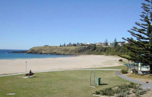 A beach view with a grassy area, a pathway, and a few buildings in the background.