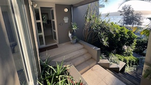 Un balcon donnant sur la mer, un escalier menant à l’entrée et diverses plantes en pot.