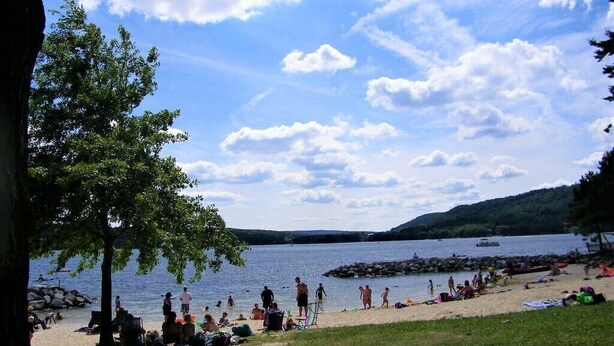Uma praia à beira do lago com pessoas relaxando e tomando sol, um barco na água e uma colina arborizada ao fundo.