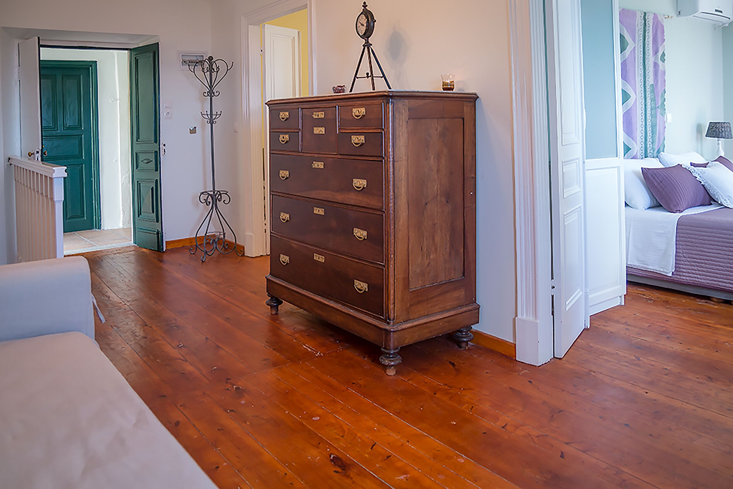 A room with wooden flooring, a dresser with multiple drawers, a bed with pillows, and a door leading to another room.