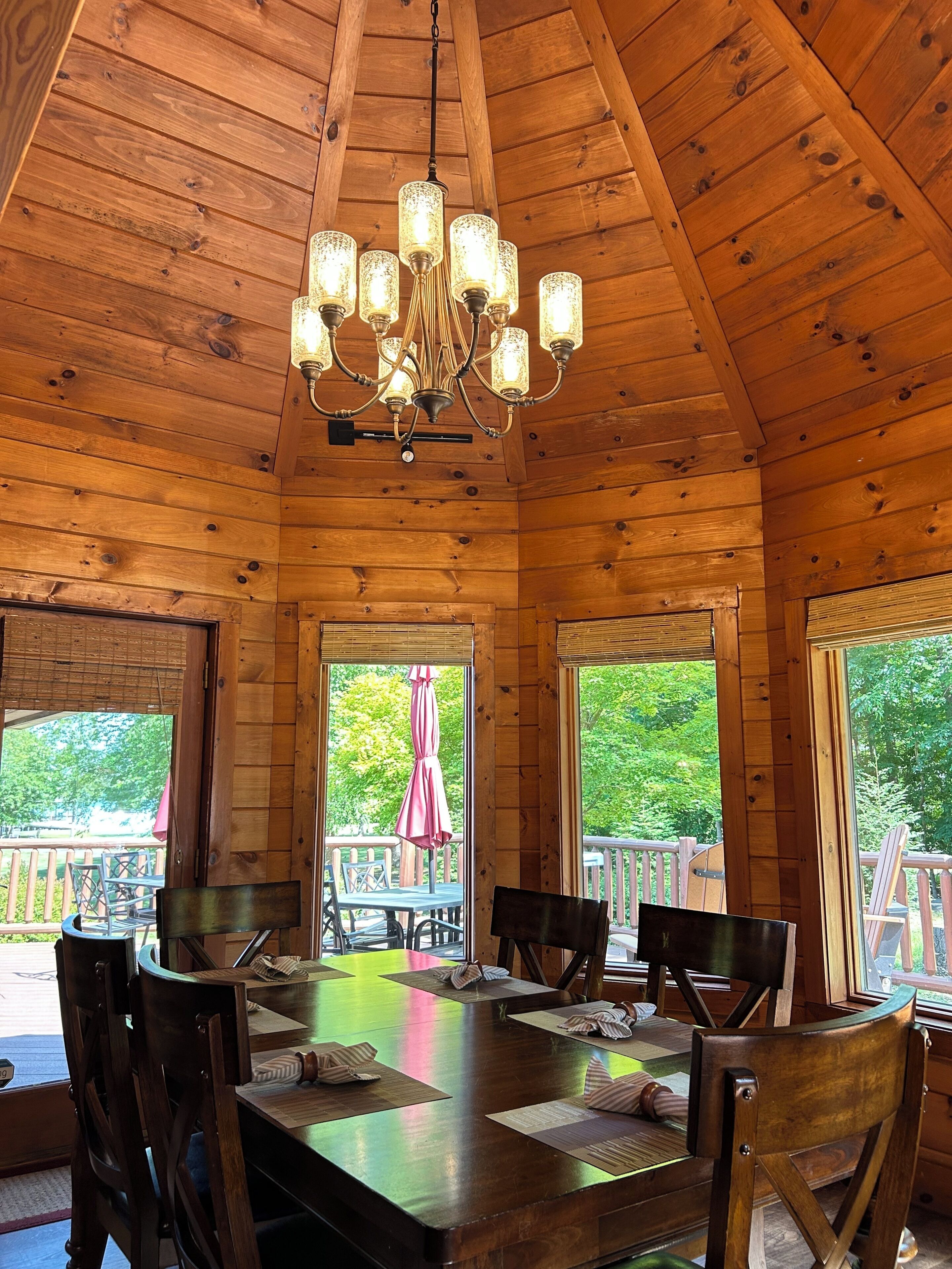 A wooden cabin with a dining area featuring a large table and chairs, a chandelier, and large windows with views of a deck and greenery.