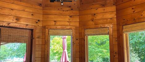 A wooden cabin with a dining area featuring a large table and chairs, a chandelier, and large windows with views of a deck and greenery.
