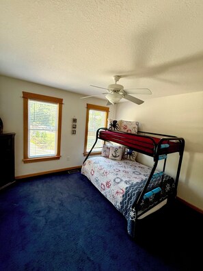 A room with a bunk bed, a ceiling fan, and two windows with blinds.
