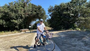 A man standing next to a bicycle with a basket, on a paved path with trees and a wooden structure in the background. - Escape to the Beech House: A Wooded Family- Friendly Retreat Close to the Beach! (Kitty Hawk)
