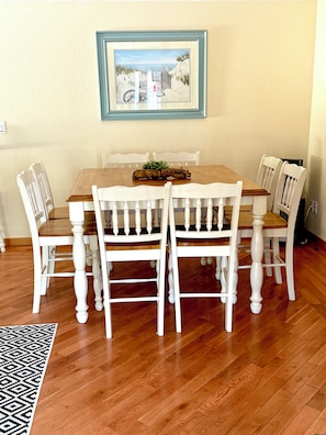 A dining area with a wooden table and white chairs, a framed picture on the wall, and a patterned rug on the floor.
