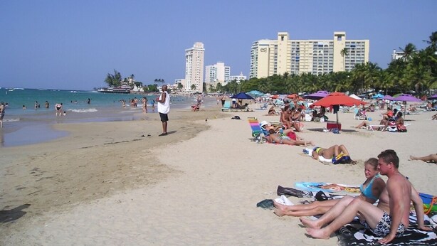 Una platja amb gent que pren el sol i nedant, palmeres i un hotel al fons.
