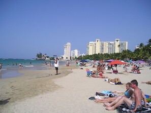 A beach with people sunbathing and swimming, palm trees, and a hotel in the background.