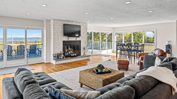 A spacious living room with a fireplace, a large sofa, and a balcony with outdoor seating. - Panoramic lake view retreat with firepit! Centrally located on Wine Trail! (Penn Yan)