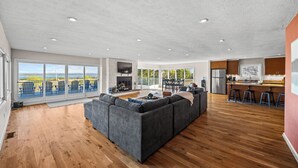 A spacious living area with a large gray sectional sofa, wooden flooring, and a fireplace. - Panoramic lake view retreat with firepit! Centrally located on Wine Trail! (Penn Yan)