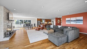 A spacious living room with a fireplace, a grey sectional sofa, a dining area, and a kitchen in the background. - Panoramic lake view retreat with firepit! Centrally located on Wine Trail! (Penn Yan)