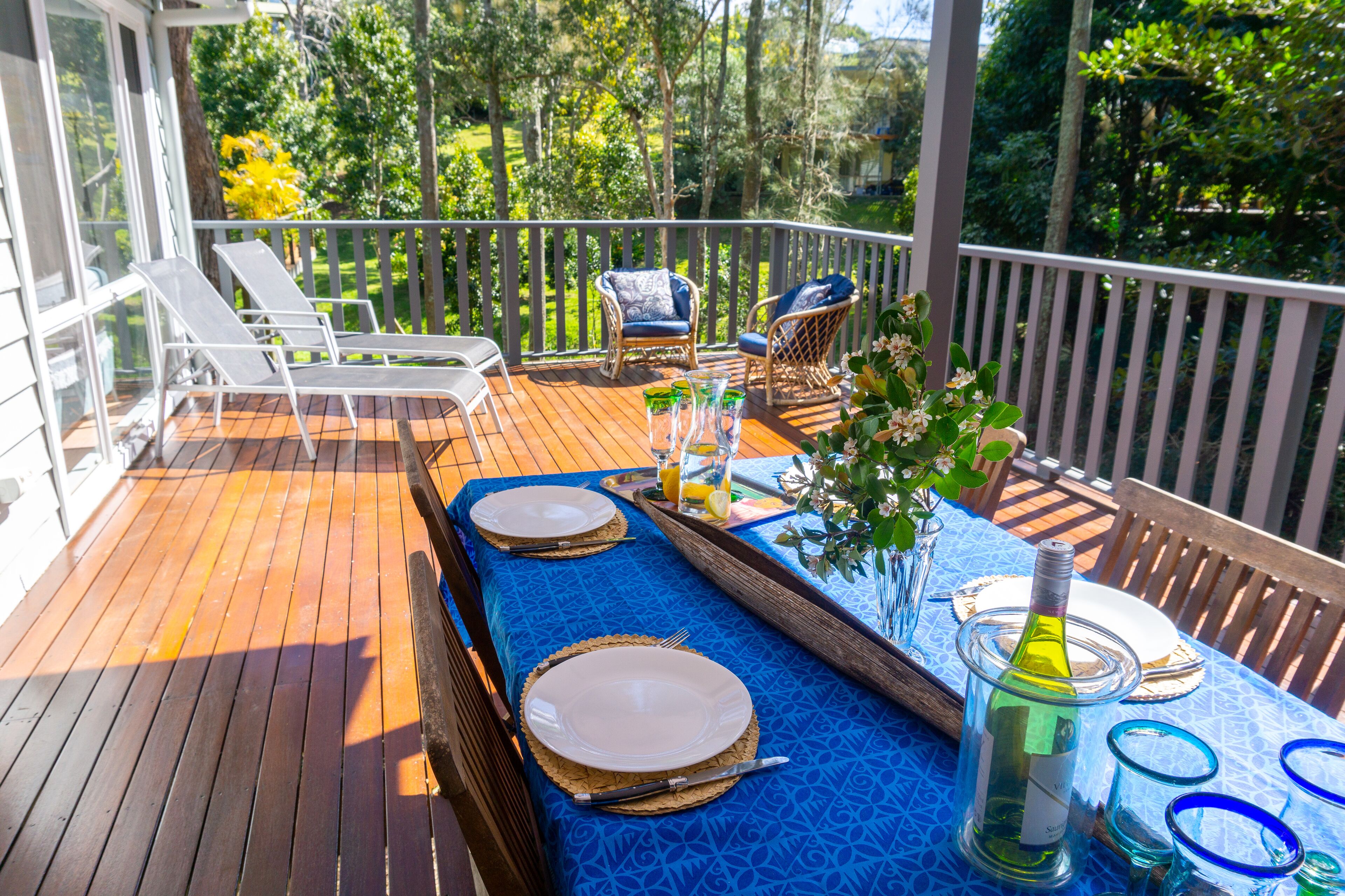 A wooden deck with a table set for two, featuring a blue tablecloth, two white plates, a wine bottle, and a glass pitcher.
