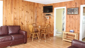 Ein Zimmer mit Holzwänden, einem braunen Ledersofa, einem Esstisch mit Stühlen, einem an der Wand befestigten Fernseher und einem kleinen Holztisch mit einem Fernseher darauf.