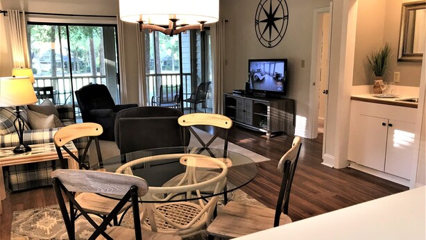 A modern living room with a large window, a dining area with a glass table, and a kitchen area with a TV. - Best of Pawleys Plantation!   Inviting Retreat for Golfers and Beach Lovers (Pawleys Island)