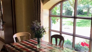 A dining area with a checkered tablecloth, two chairs, a vase with flowers, and a large window with a view of greenery. -  Charming old-world country cottage, near Adare  (Kildimo)