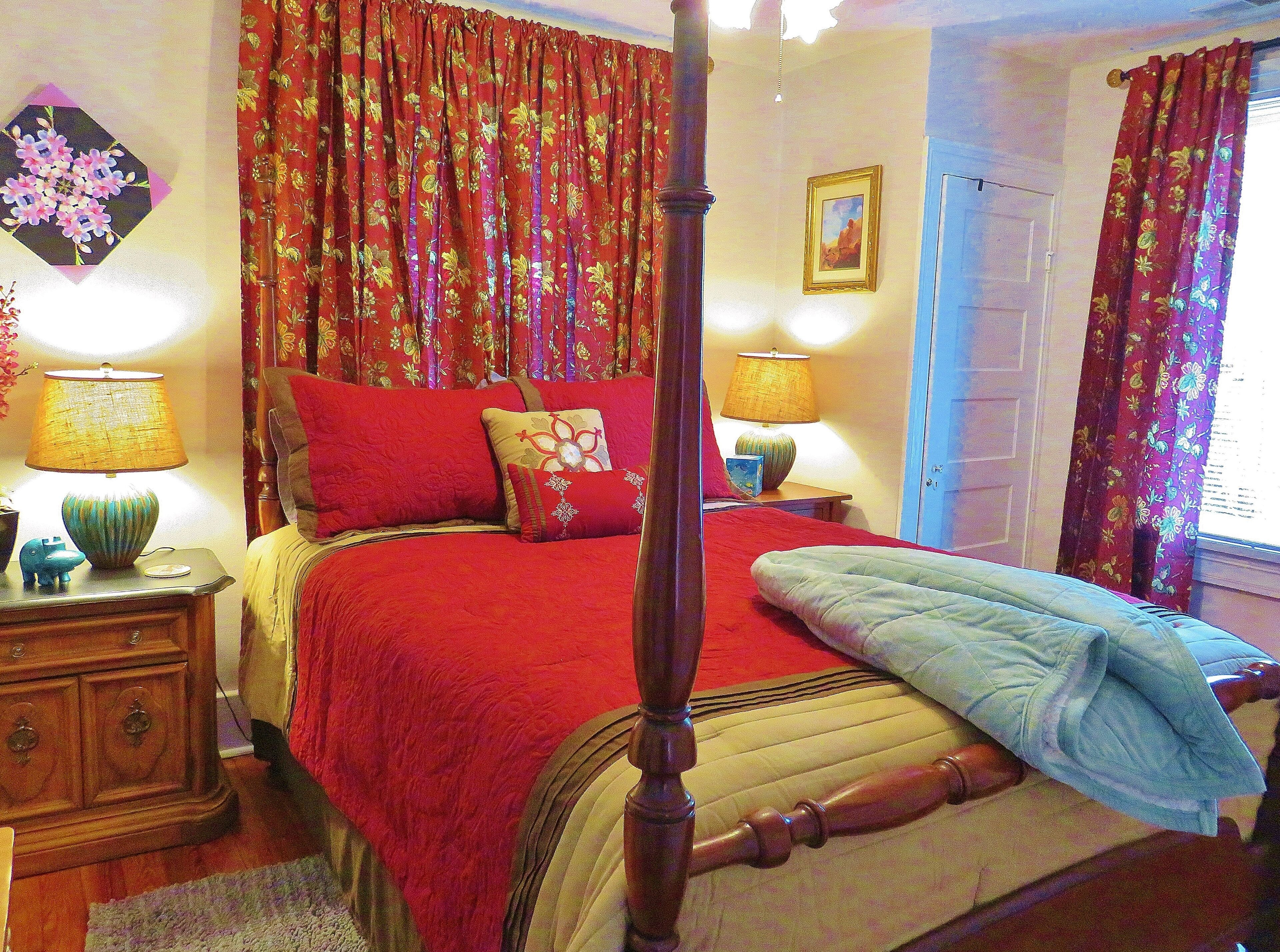 Un lit à baldaquin avec une literie rouge et beige, une table de chevet en bois avec une lampe et des rideaux à motifs floraux.